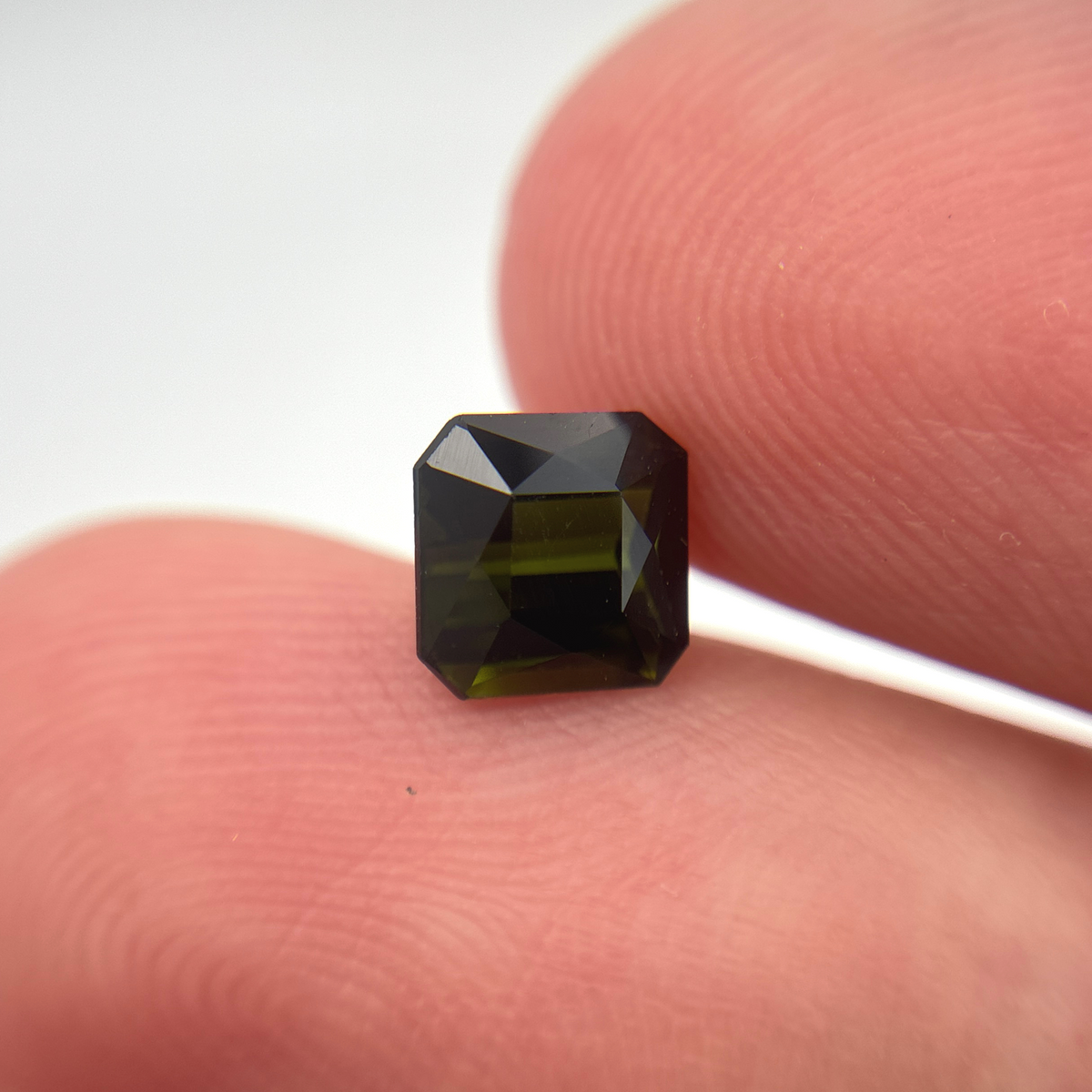 A small rectangular square faceted dark green tourmaline held between two fingers to show its size, photographed against a plain light background.