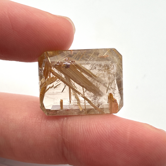 A rectangular quartz with golden rutile needle-like inclusions, held between two fingers against a plain white background.