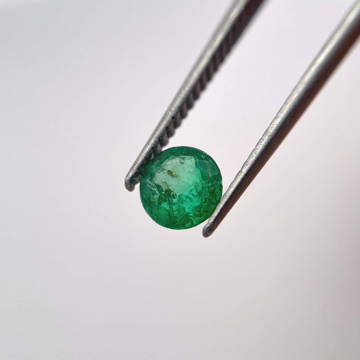 A round, faceted green emerald held between silver metal tweezers against a clean, white background.