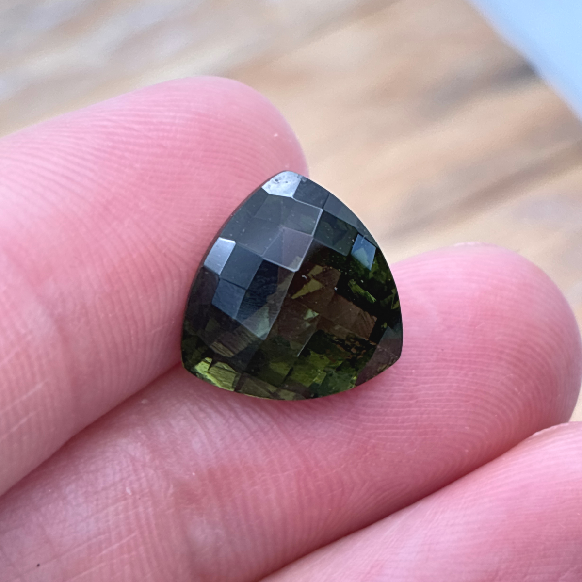 A faceted triangular dark olive green Moldavite and black coloration, held on a person's fingers against a blurred wooden background.
