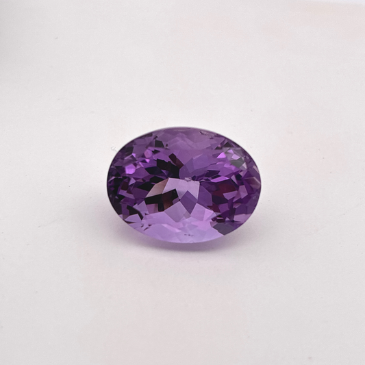 An oval-cut purple amethyst with faceted cuts,  and a chip on the back, displayed on a clean white background.