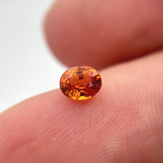 A small oval-cut orange sapphire resting on a person's fingertips against a soft white background.